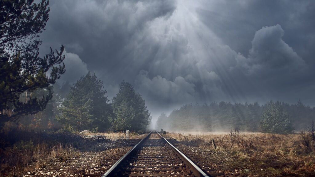 Railroad straights heading straight off into the distance, through a pine forest, under storm skies.  Sunbeams race down through the clouds.