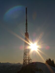 A brilliant sun shining behind a radio tower.